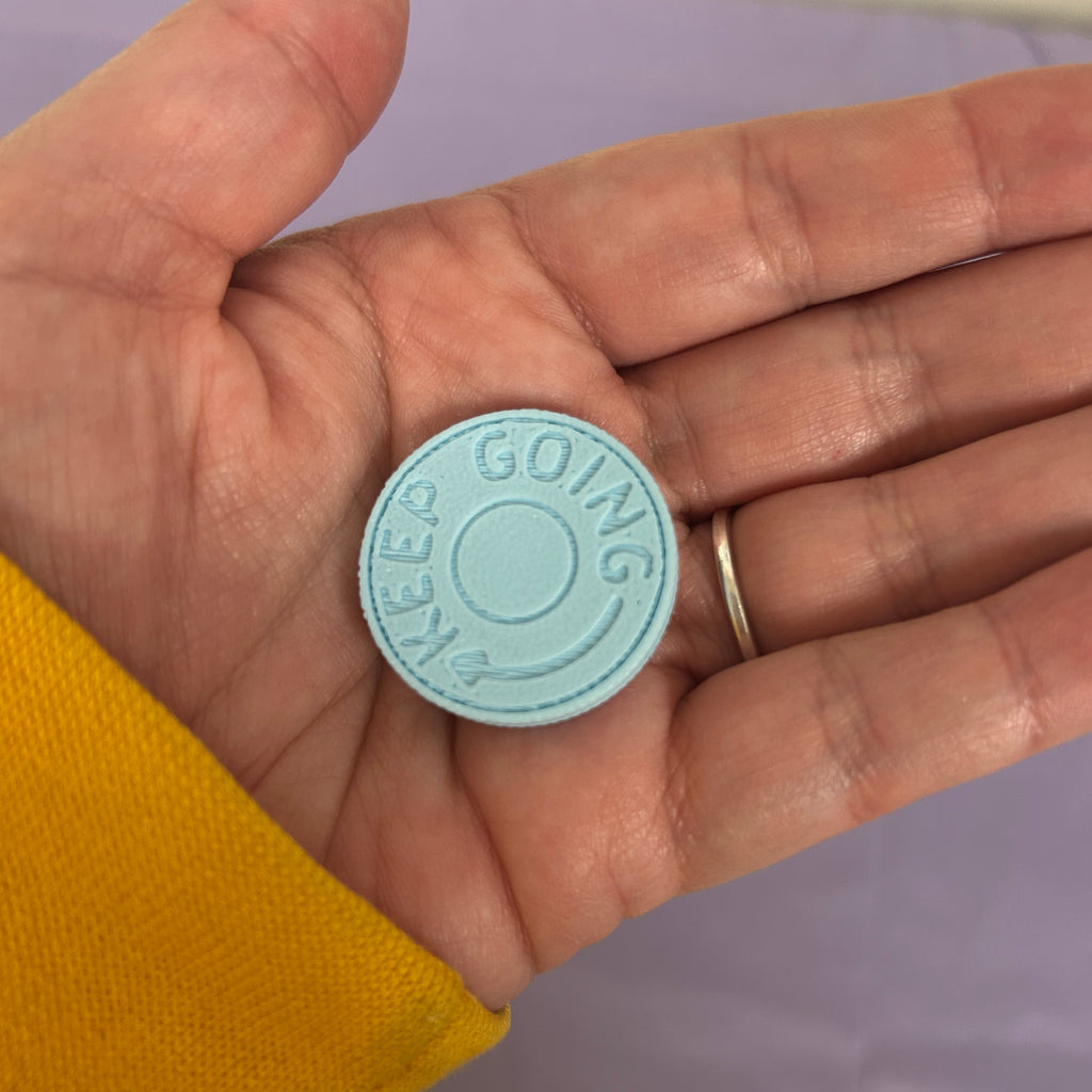 A small round light blue button with the words 'KEEP GOING' debossed around the edge, held in a person's hand wearing a yellow sleeve and a gold ring.