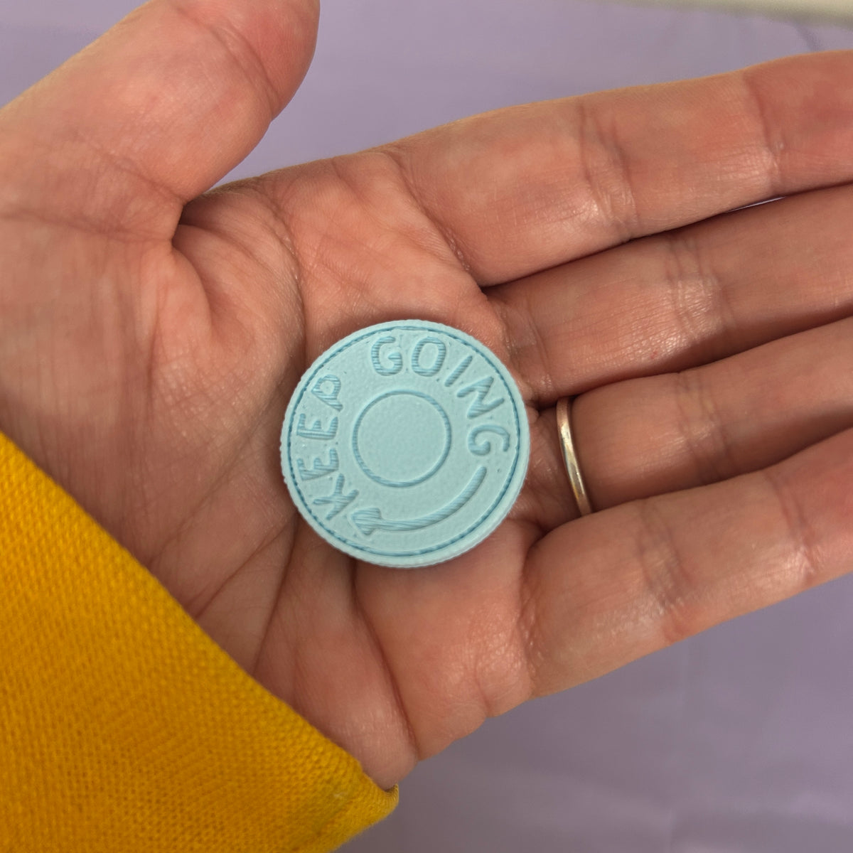A small round light blue button with the words 'KEEP GOING' debossed around the edge, held in a person's hand wearing a yellow sleeve and a gold ring.