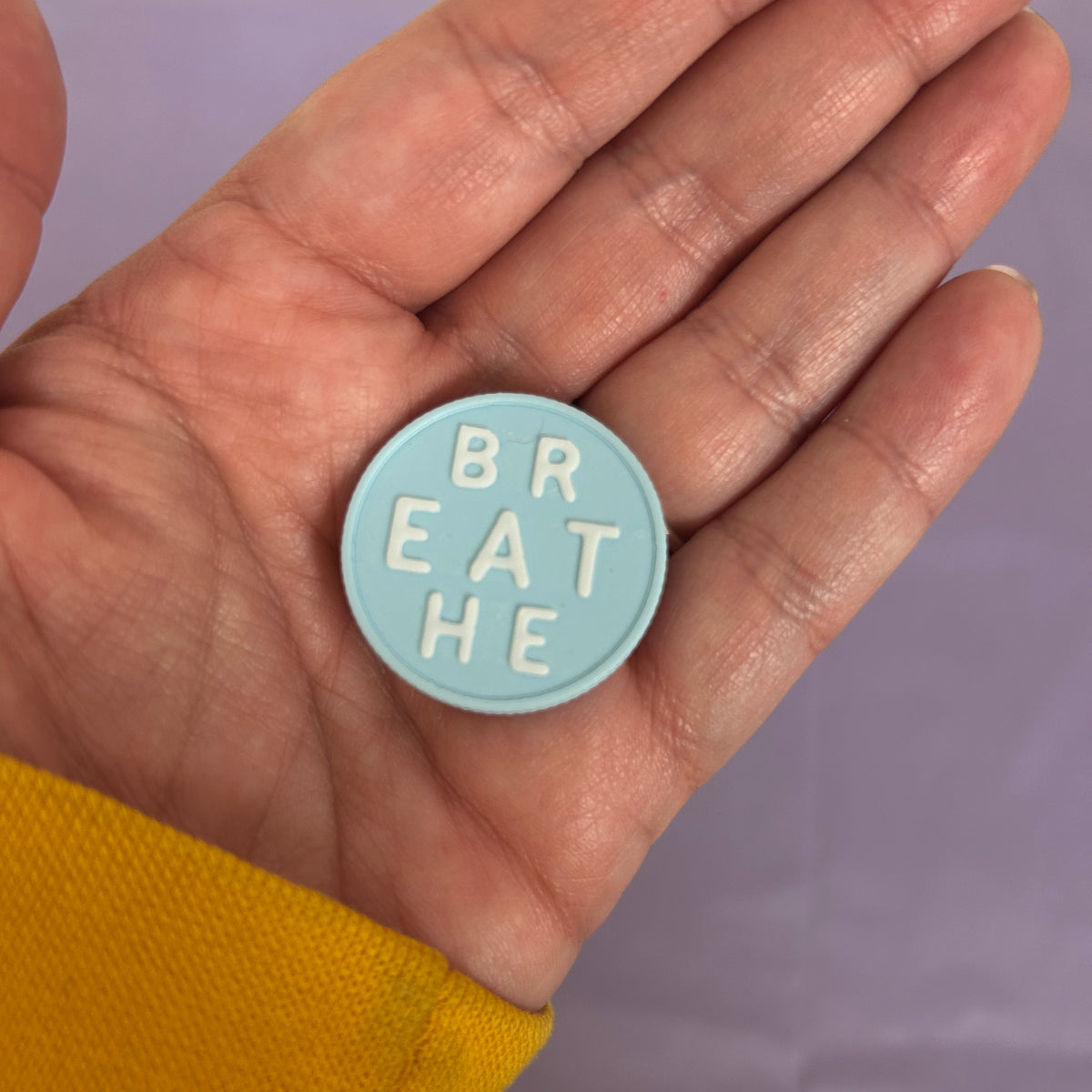 A small round light blue pin or badge with the words 'BREATHE' in raised white letters, held in an open hand against a soft lavender background.