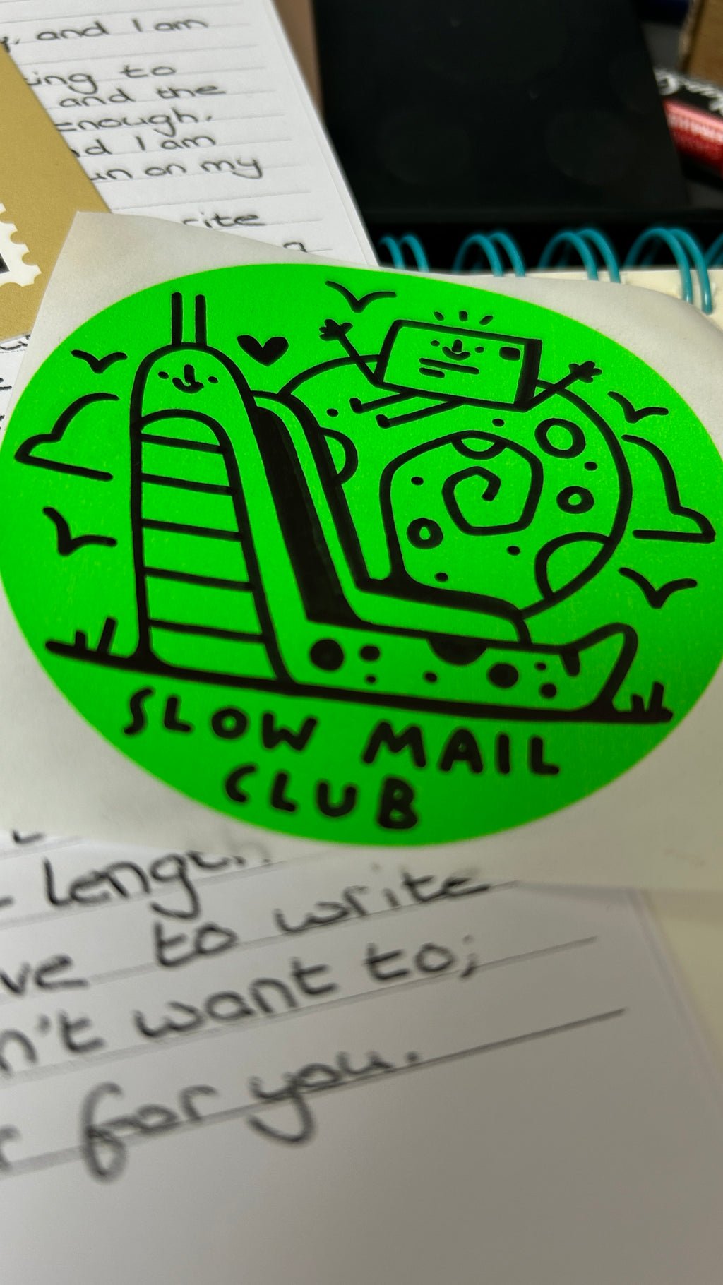 Green sticker with 'Slow Mail Club' design on a surface with handwritten text.