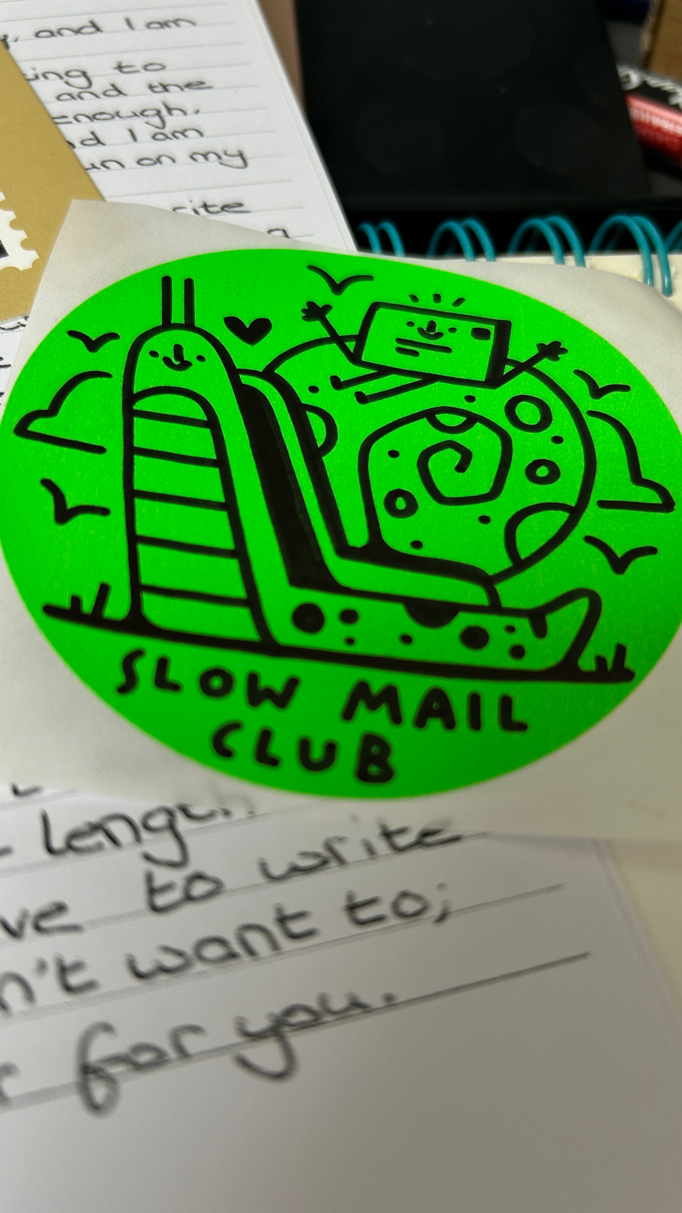 Green sticker with 'Slow Mail Club' design on a surface with handwritten text.
