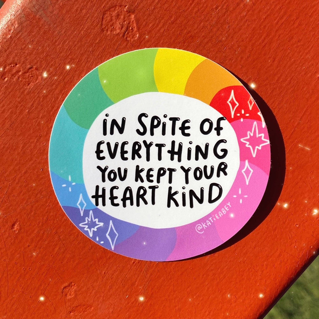 A round sticker with a rainbow border and the handwritten text 'IN SPITE OF EVERYTHING YOU KEPT YOUR HEART KIND' in black, placed on a textured orange-red surface.