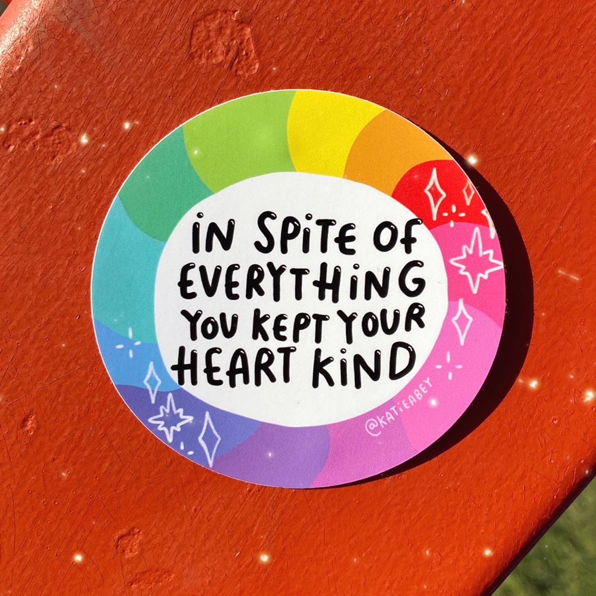 A round sticker with a rainbow border and the handwritten text 'IN SPITE OF EVERYTHING YOU KEPT YOUR HEART KIND' in black, placed on a textured orange-red surface.