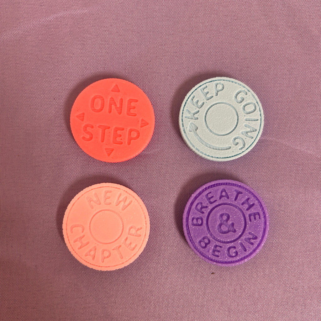 Four circular motivational or mindfulness tokens arranged in a 2x2 grid on a textured mauve fabric background. The top-left token is coral orange with the words 'ONE STEP' and small triangles.