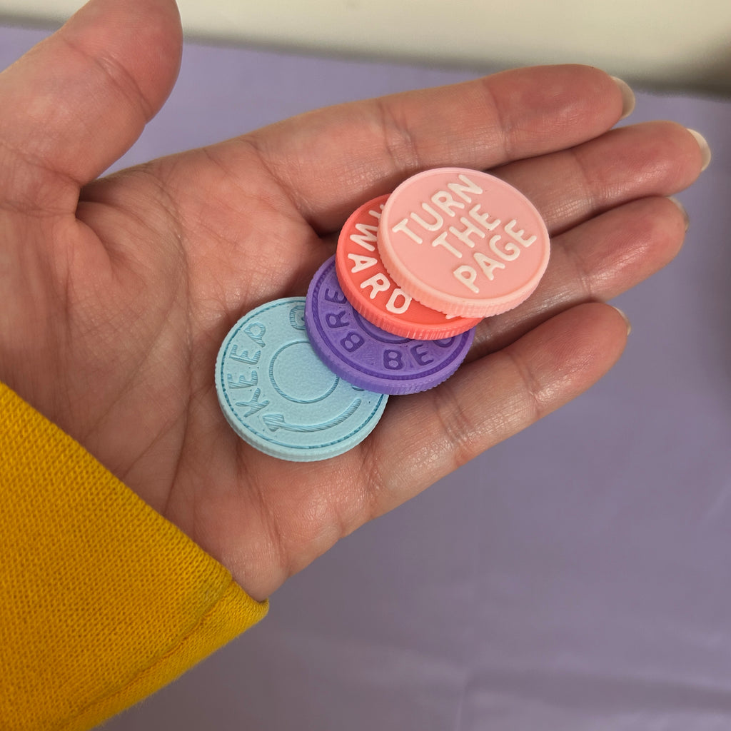 Four small round pastel buttons or badges held in a person's hand. The buttons are light blue, purple, coral orange, and pink, with white text reading 'TURN THE PAGE', 'AM AR', '38 GRA', and 'SWEEES'.